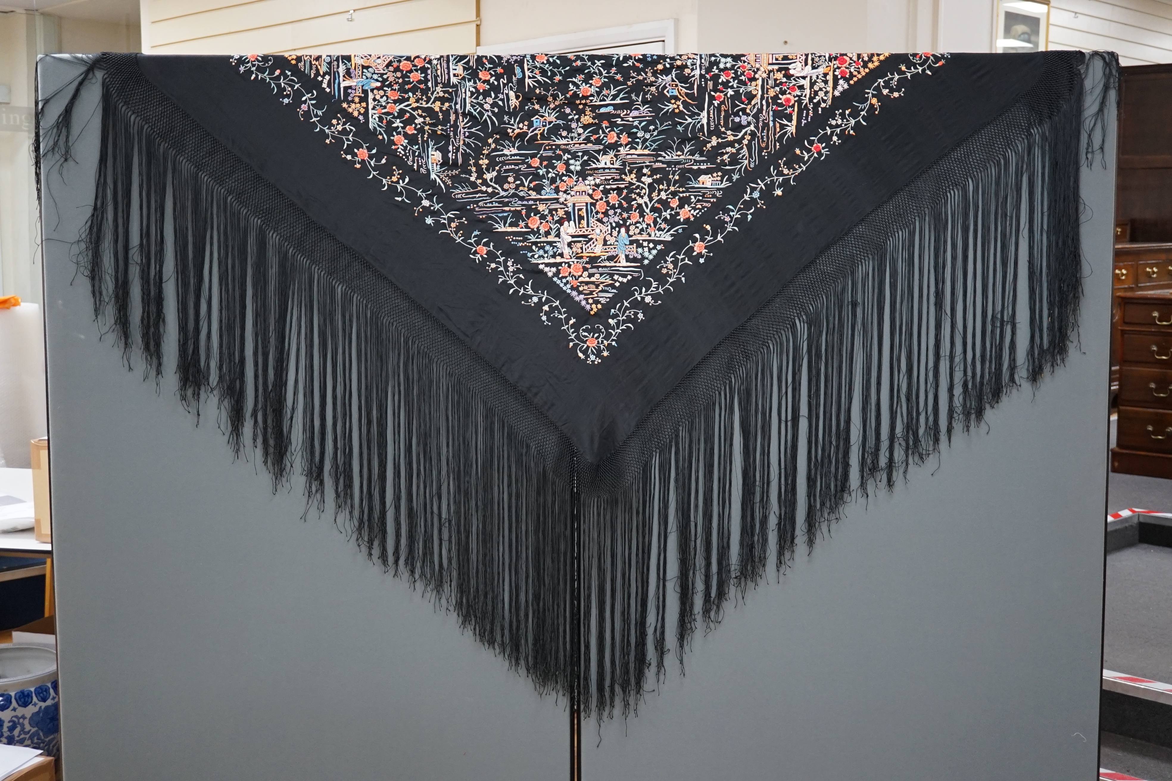 An early 20th century Chinese black silk shawl, with all over coloured floral embroidery, embroidered with multi-coloured silks and a long silk fringe, unusually the four corners are embroidered with a figurative pagoda